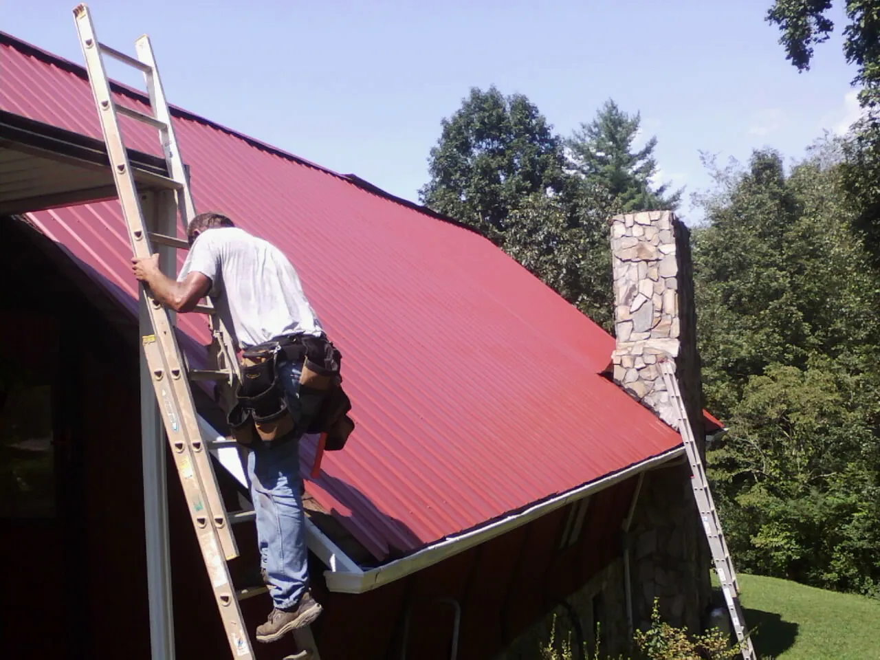 Roofer on a ladder beside a red metal roof and stone chimney — Soffit & Fascia Repair by Roofing Brothers in Portersville